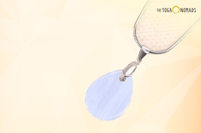 A pendant with a blue lace agate stone attached to a silver-colored chain. The stone has a characteristic light blue color with white banding patterns, typical of blue lace agate. The background is pale yellow with soft white gradients.