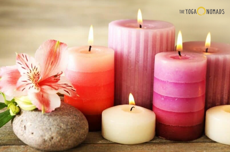 A collection of six lit candles of various sizes and colors, with a pink flower resting on a stone next to them. The candles are arranged on a wooden surface, suggesting a peaceful and serene setting, possibly for meditation or relaxation.