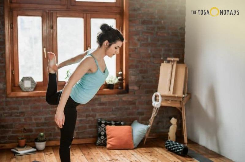 An individual practicing yoga in a room with a rustic aesthetic. The person is standing on one leg, with the other leg bent and held up by their hand, suggesting a balancing pose. The setting includes wooden floors, a large window with wooden frames, and various items like cushions and a rolled-up yoga mat.