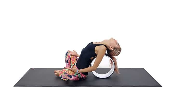 An individual using a yoga wheel to assist in a backbend pose on a yoga mat. The person is wearing a black tank top and patterned leggings. The yoga wheel helps enhance flexibility, maintain proper alignment, and deepen the stretch in various yoga poses.