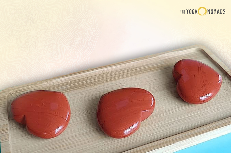 Three polished red jasper stones placed on a wooden tray. The stones have a deep, rich red color and are smooth with an oval shape, each having unique contours. The background is a light blue surface with a subtle floral pattern.