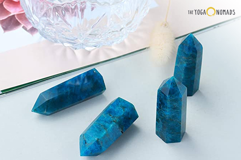 A collection of five blue apatite crystal towers arranged in a line on a reflective surface, with the tallest in the center. The background includes a blurred object that appears to be a round crystal bowl and some plant material.