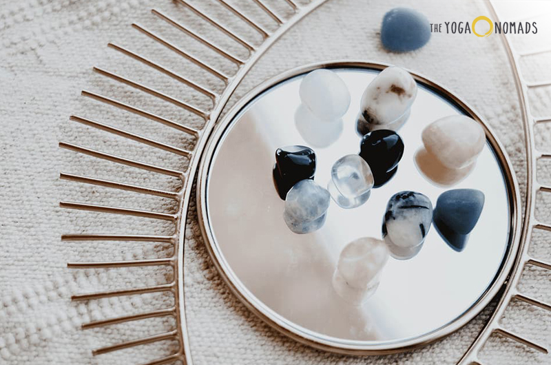 A collection of smooth, polished stones resting on a round mirror these stones in this context represent the third eye chakra crystals. Part of a beige textured surface and the edge of a circular object with radiating lines are visible in the background.