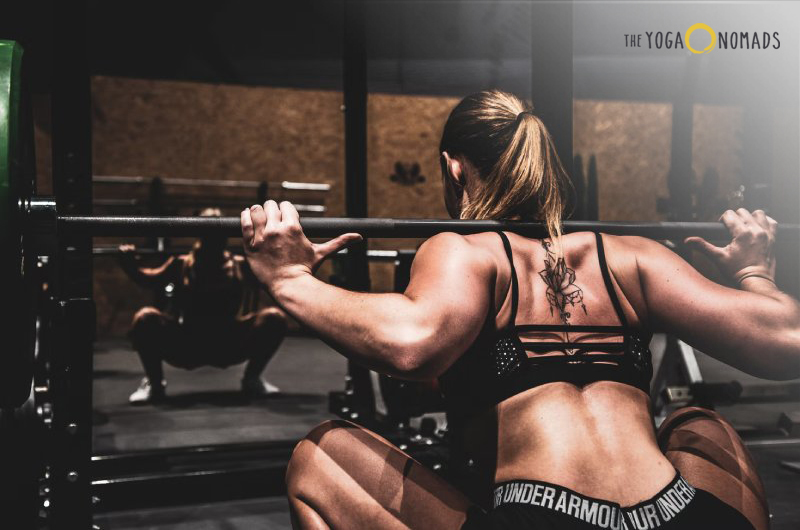 An individual is in a gym setting, preparing to perform a squat with a barbell on their shoulders. The person has visible muscle definition, indicating strength and fitness. The background includes weightlifting equipment, suggesting a focus on physical training.