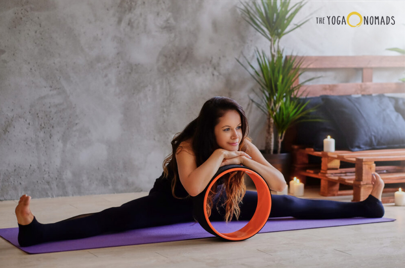 An individual performing a yoga pose on a purple yoga mat. The person is seated with legs spread wide apart in a straddle position and is leaning forward, holding a yoga wheel with both hands. The setting appears to be an indoor space with a calm and serene ambiance, indicated by the presence of candles and a plant in the background.