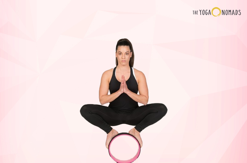 An individual in the Lotus position with hands pressed together in a prayer-like gesture known as Anjali Mudra. The person is seated on a pink yoga wheel, with a light pink gradient background.