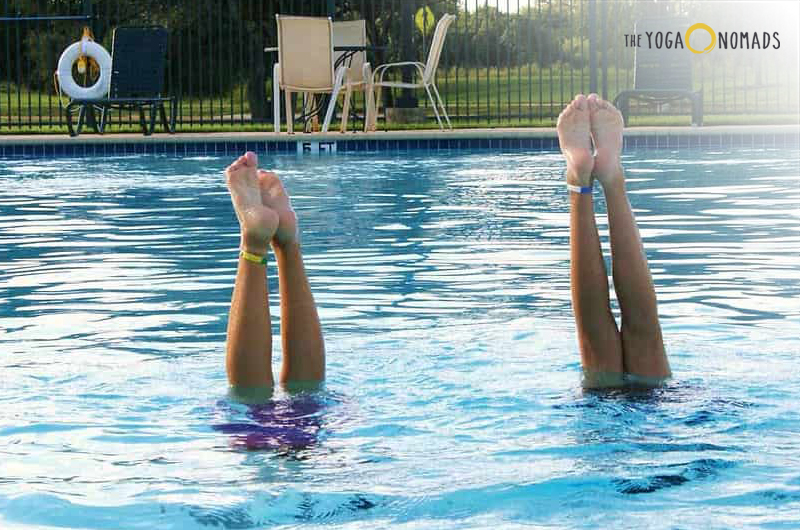 The lower half of two individuals submerged in a swimming pool with only their legs and feet visible above the water. The individuals appear to be doing an upside-down yoga or exercise pose. In the background, there are chairs and a table near the poolside, indicating a leisure or recreational setting. The sky is visible and appears to be during daylight hours