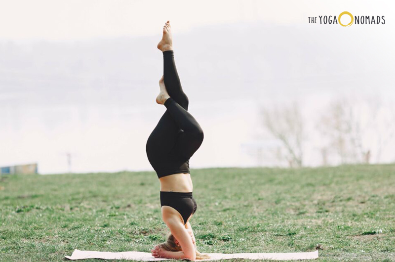 An individual is performing a yoga pose outdoors on a mat, with legs lifted upwards and arms supporting the body. The background includes grass and a blurred view of what appears to be a body of water.