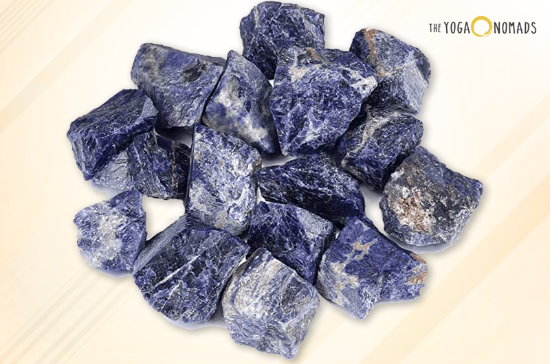 A collection of sodalite stones against a light background with a subtle diagonal stripe pattern. Sodalite is a rich royal blue tectosilicate mineral with deep blue color and white calcite interspersed. The stones are unpolished, showing their natural rough texture and angular shapes.