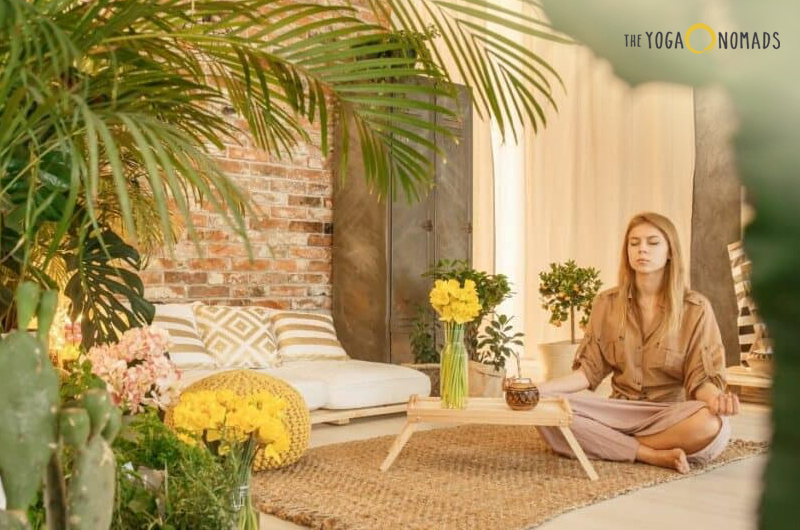 An individual sitting cross-legged on a woven mat in a serene indoor setting. Surrounding the individual are potted plants, including large palm leaves, flowers in vases, and soft furnishings like cushions and a low table. A brick wall adds texture to the background.