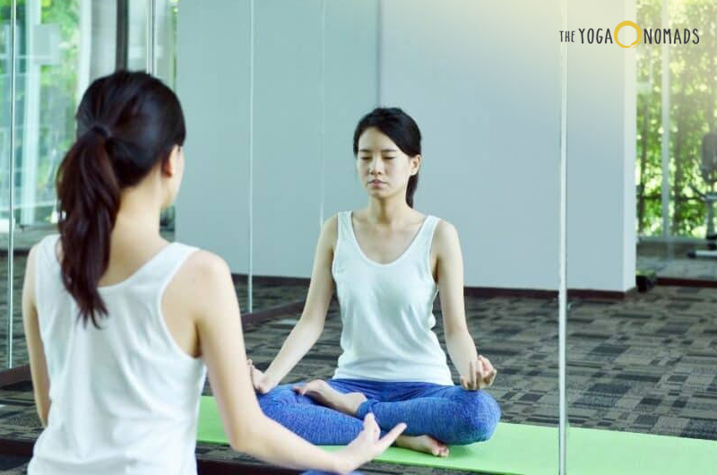 The image features an individual seated on a green yoga mat in a meditative pose, facing a mirror that reflects their image. Large glass windows in the background reveal trees and daylight, creating a serene and peaceful atmosphere ideal for yoga or meditation.