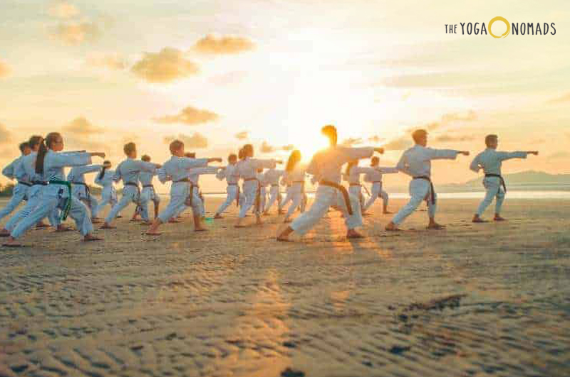 A group of individuals practicing martial arts on a beach at sunset or sunrise. The individuals are in a staggered formation, performing a synchronized move with one arm extended forward. The sun is low on the horizon, casting a warm glow and long shadows on the sand. The sky is partly cloudy.