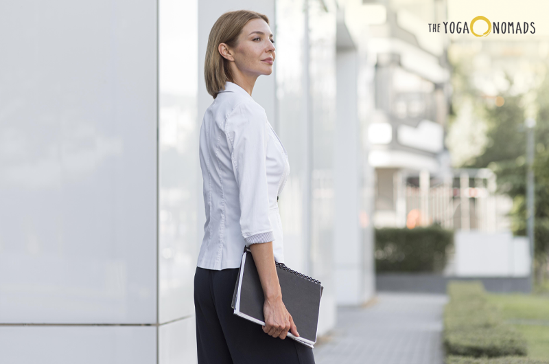 An individual is standing outdoors, holding a black notebook or folder. They are dressed in a white shirt with three-quarter sleeves and dark pants. The background includes a modern urban setting with buildings and greenery. The logo "THE YOGA NOMADS" is visible in the top right corner.